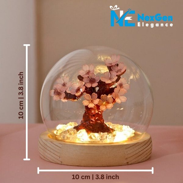 A close-up of a small, decorative cherry blossom tree inside a clear glass dome with its dimensions displayed. The tree has light pink flowers on dark branches and sits on a light wood base containing clear, lit crystals. White lines with double arrows indicate that the dome is 10 cm or 3.8 inches tall and 10 cm or 3.8 inches wide. The logo "NexGen Elegance" is in the top right corner.