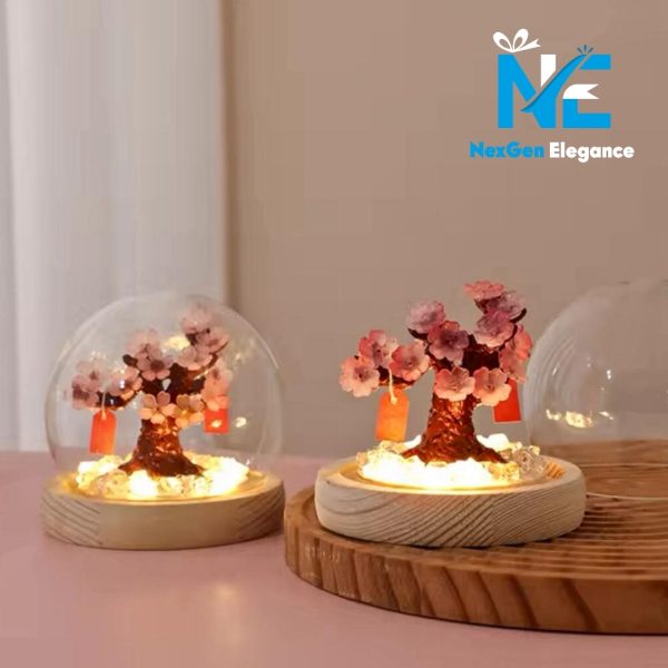 A wider shot shows two decorative cherry blossom trees, each with pink flowers, on a pink surface. The tree on the left is enclosed in a glass dome, while the one on the right is not, showing a clear, empty glass dome next to it. Both trees have small, red paper tags hanging from their branches and are lit from below, illuminating the clear crystals on their light wood bases. The logo "NexGen Elegance" is in the top right corner.