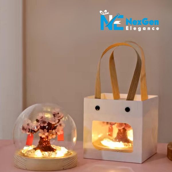 A decorative cherry blossom tree inside a glass dome, with red tags hanging from its branches, sits on a pink surface next to a white gift bag with a cutout window. The tree is lit from below and the light shines through the window of the gift bag, which has brown handles and two small black circles resembling eyes. The logo "NexGen Elegance" is in the top right corner.