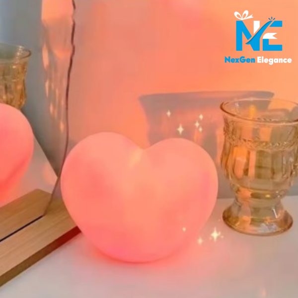 Glowing 3D heart lamp emitting soft pink light on a white surface with decorative glass cups and mirror reflection, NexGen Elegance logo
